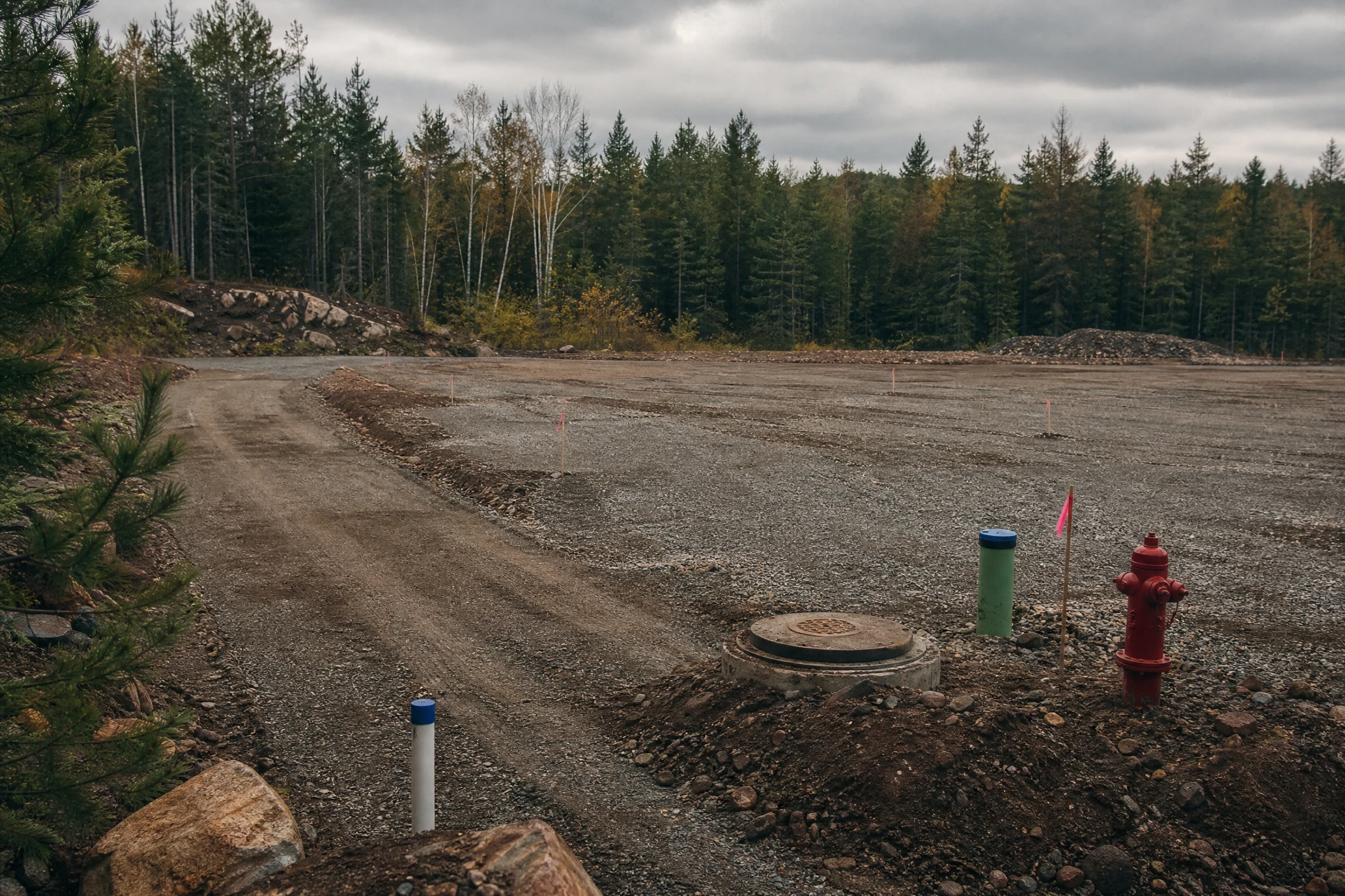 A cleared gravel pad site with fresh utility stub-outs, a fire hydrant, and survey flagging at the edge of a small Minnesota town, forest tree line beyond