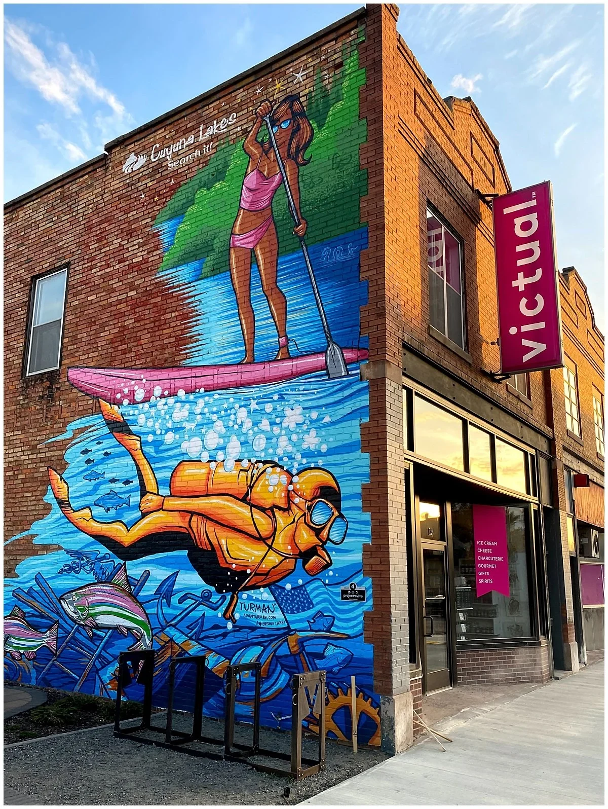 A photograph of a colorful mural of a paddleboarder and a scuba diver on the side of a downtown Crosby storefront, with the Victual shop sign and a bike rack out front
